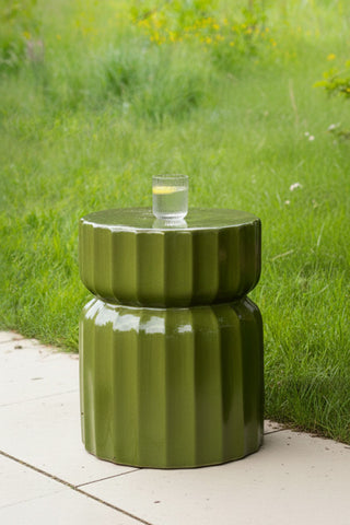 The Ribbed Edge Round Side Table in Green Glaze styled with a glass tumbler on, displayed outdoors with grass in the background.