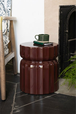 The Ribbed Edge Round Side Table in Rust Glaze styled with a mug and book on, displayed next to a chair and a plant.