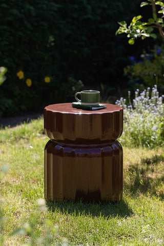 The Ribbed Edge Round Side Table in Rust Glaze displayed with a mug and book on, outside on some grass.