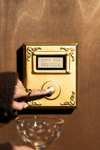 The Ring For Refills Gold Wall Ornament shown against a wood wall with someone holding up a champagne glass and pressing the button. 