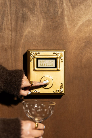 The Ring For Refills Gold Wall Ornament shown styled on a wood textured wall with someone pressing the button and holding up a champagne glass. 