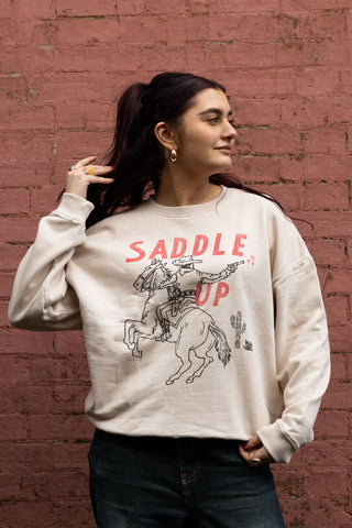 The Saddle Up Cowboy Sweatshirt in Oat worn by a model standing in front of a brick wall.