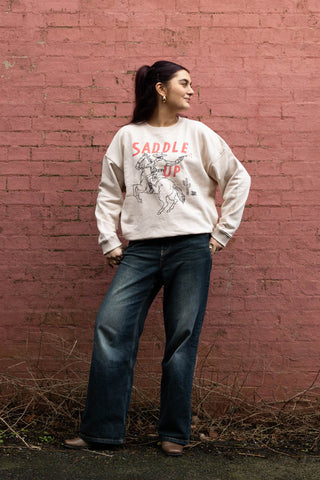 The Saddle Up Cowboy Sweatshirt in Oat worn by a model, posing in front of a brick wall.