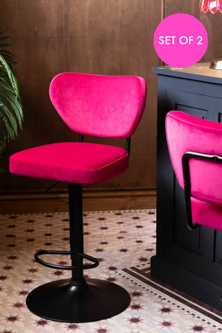 The Set Of 2 Adjustable Bar Stools in Harry's Hot Pink shown against a black counter with dark wood walls in the background. 