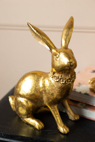 One of the Set Of 2 Beautiful Gold Rabbit Ornaments styled on a sideboard with some books in the background.