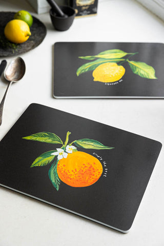 Lifestyle image of the Set Of 2 Orange & Lemon Placemats displayed with fruit and kitchen accessories on a white window ledge.