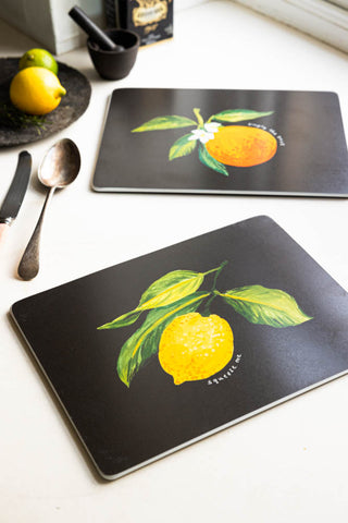 Lifestyle image of the Set Of 2 Orange & Lemon Placemats displayed with fruit, cutlery and other kitchen accessories. 