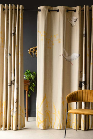 The Set Of 2 Natural Cranes Lined Curtains, with one curtain pulled across more, styled with a chair, table, plant and bird wall decoration.