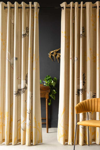 The Set Of 2 Natural Cranes Lined Curtains hanging from a black rail, with a yellow chair in front and a table, plant and bird wall decorations behind.