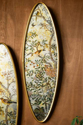 One of the Set of 2 Framed Oval Chinoiserie Wall Hangings shown on a wood textured wall, with birds, trees and flowers in the design. 