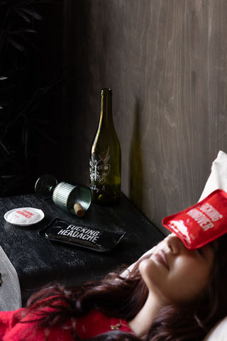 Person lying down with a red cooling/heating pad on their forehead, next to a bottle, glass and another pad labeled 'Fucking Headache'.
