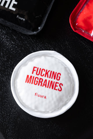 Round white cooling/heating pad with the word 'Fucking Migraines' on, displayed on a dark surface.