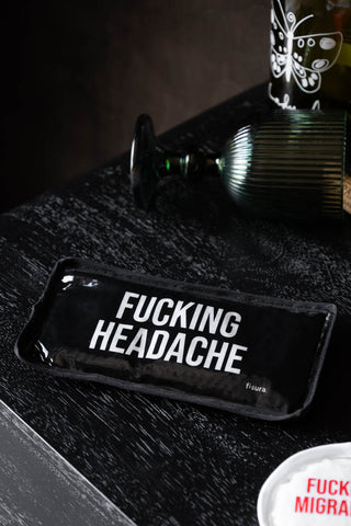 A black cooling/heating pad with the words 'Fucking Headache' on, styled on a bedside table with another pad, a wine bottle and a wine glass on its side.
