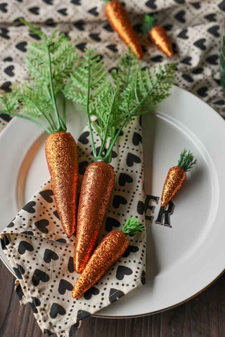 Lifestyle image of the Set Of 9 Glitter Carrot Decorations displayed on a Le Diner Bistro Dinner Plate, with a Monochrome Heart Cotton Napkin and Monochrome Heart Cotton Tablecloth on the table beneath. 