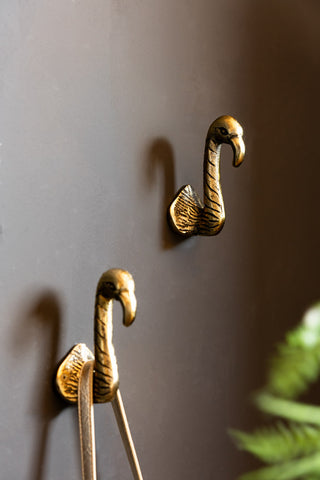 The Set of 2 Antique Gold Flamingo Head Coat Hooks shown against a dark wall with a palm in the background. 