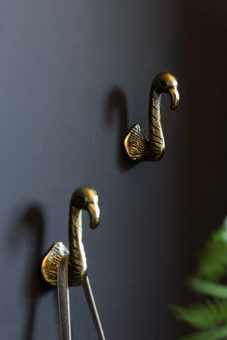 The Set of 2 Antique Gold Flamingo Head Coat Hooks shown against a dark wall with a palm in the background. 