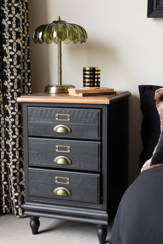 Black and wood bedside table styled with a lamp, books and candle on, next to a curtain and a bed.
