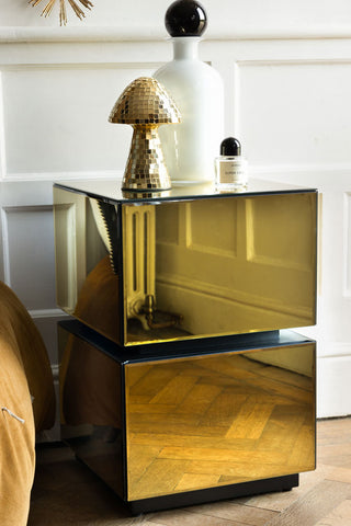 Gold mirrored bedside table with a gold mushroom ornament, perfume and white bottle styled on top, in front of a white wall.
