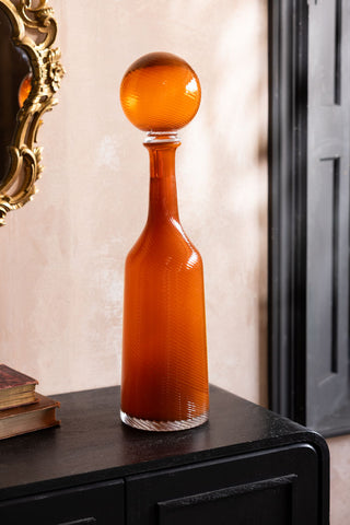 The Seventies Swirl Orange Glass Decanter shown against a plaster wall on a black sideboards. 
