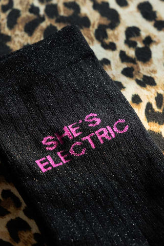 The She's Electric Cotton Glitter Socks shown against a leopard background. Socks are black with hot pink text that says 'she's electric'.