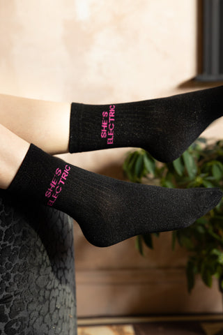 The She's Electric Cotton Glitter Socks shown with the model's legs hanging over a sofa against plaster walls. 