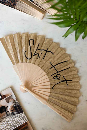 The Shit Hot Black & Gold Glitter Fan displayed on a marble table other fans, a magazine and a plant.