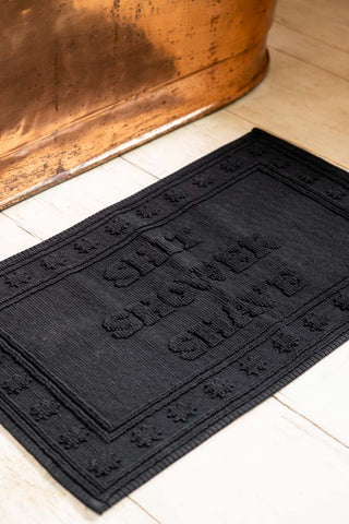 The Shit Shower Shave Black Bath Mat shown next to a copper bath. 