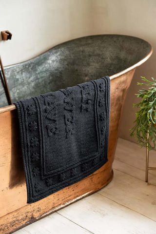 The Shit Shower Shave Black Bath Mat shown draped over the side of a copper bath. 