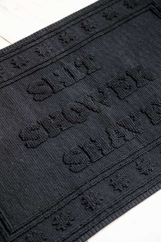 A detail crop showing the texture and slogan on the Shit Shower Shave Black Bath Mat.