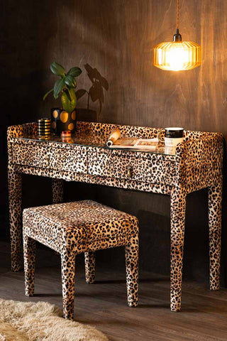 The Signature Leopard Print Velvet Dressing Table & Stool is shown against a textured wood wall with plants and accessories styled on the glass top. 