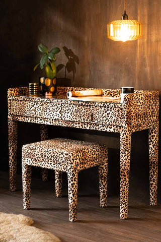 A side angle of the Signature Leopard Print Velvet Dressing Table & Stool, shown against a dark textured wood wall with an amber ceiling light.