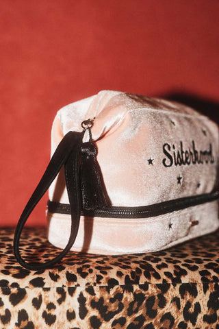 The side of the Sisterhood Blush Pink Velvet Washbag showing the black handle and tassel zip tie, with the pale pink velvet washbag sat on a leopard surface against brick red walls.