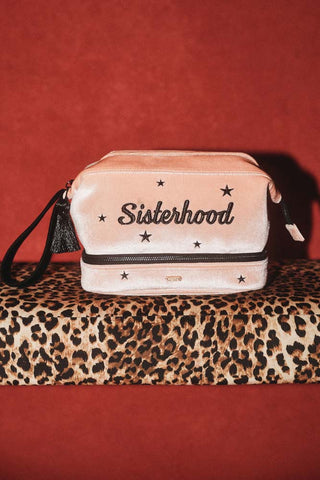 The Sisterhood Blush Pink Velvet Washbag shown sat on a leopard print bench against brick red walls. 