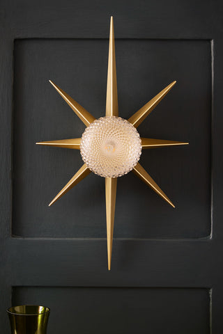 Lifestyle image of the Gold Starburst Wall Light displayed on a black panelled wall. 