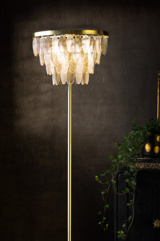 The Stunning Art Deco Glass Feather Floor Lamp switched on and displayed in front of a dark wall, next to a plant on a black sideboard.