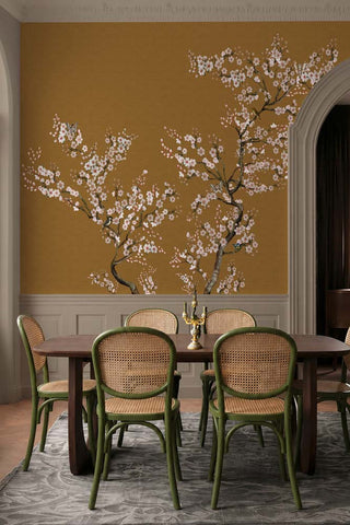The Blossom Mischief Mural Wallpaper in Antique Gold shown in a dining room in front of green rattan dining chairs. 