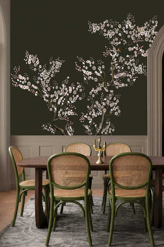 The Blossom Mischief Mural Wallpaper in Forest shown in a dining room with green rattan chairs. 