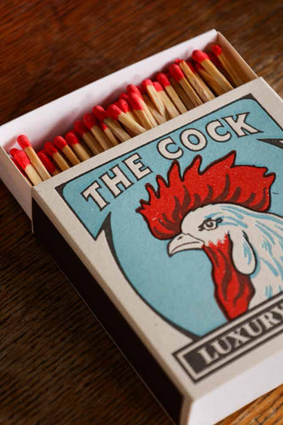 Lifestyle image of The Cock Luxury Matches displayed on a wooden table. 