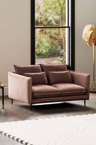 The Lola 2 Seater Sofa in Luxe Pink Velvet styled witha rug, side table and floor lamp, in front of a window.