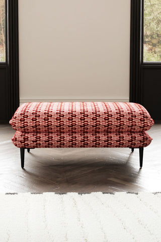 The Lola Footstool in Woven Red & Pink Lips Velvet styled on a wooden floor in front of a rug, with some windows in the background.
