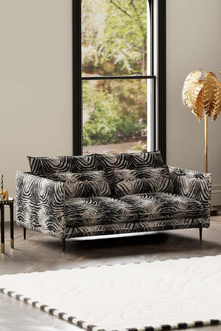 The Lola 3 Seater Sofa in Zebra Print Velvet styled with a rug, floor lamp and side table, in front of a window.