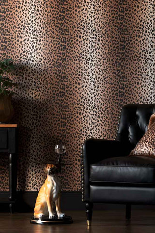 A close up of The Wild At Heart Wallpaper in Tan shown in a living room, with a black faux leather chesterfield armchair and a dog side table. 