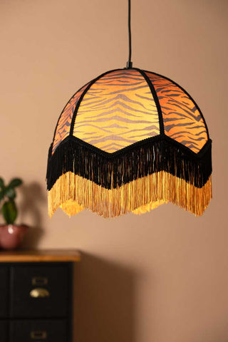 A fringed tiger print lampshade hanging in front of a plain wall, sideboard and plant.