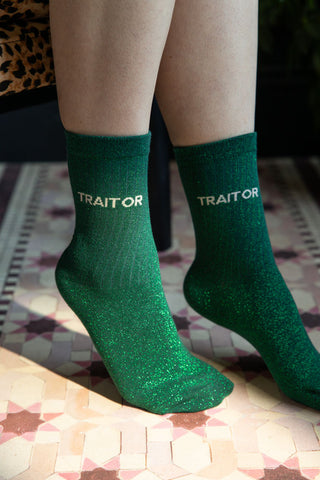 The Traitor Green Glitter Socks shown worn with feet on a pink tile-style floor. 