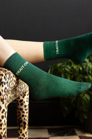 The Traitor Green Glitter Socks shown worn with legs hanging off the edge of a leopard print footstool, against a black wall. 
