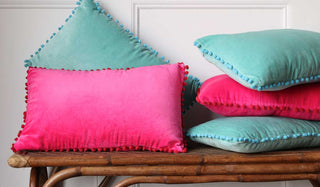 landscape image of the hot pink and pale blue pom pom cushions stacked on a bamboo wood bench