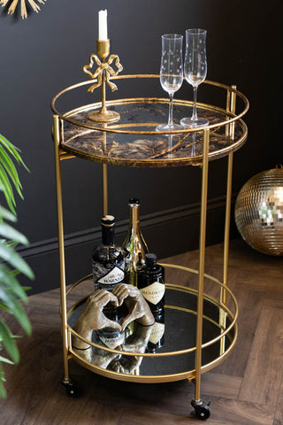 The Vintage Palms Gold Mirrored Drinks Trolley shown against a black wall with gold accessories and wine glasses styled on top. 