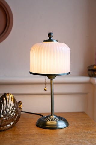 The Vintage Pink Fluted Glass Table Lamp shown with the light on on top of a wood table top and neutral panelled walls. 