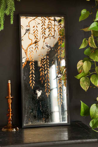 Lifestyle image of the Weeping Willow Tree & Bird Mirror styled on a black sideboard with plants and a candlestick holder. 