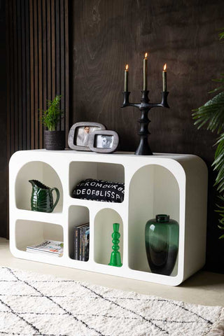 Lifestyle image of the White Alcove Shelf displayed with various home accessories inside and on the top, in front of a dark wooden wall, next to a plant and rug.